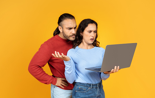 Beautiful angry millennial couple using modern laptop together, man and woman posing on yellow studio background, surfing on Internet, gambling online, copy space. Software error