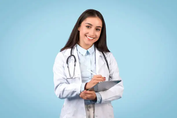 Attentive young female doctor in a white coat using digital tablet, representing thorough patient care and medical documentation, smiling at camera