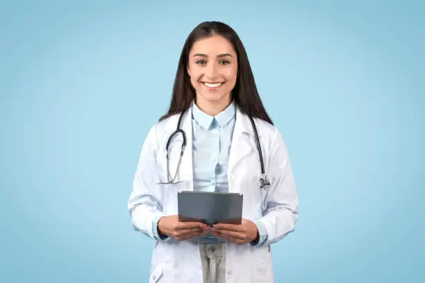 European female doctor in white coat, holding digital tablet and smiling at camera, symbolizing modern healthcare, posing against blue backdrop