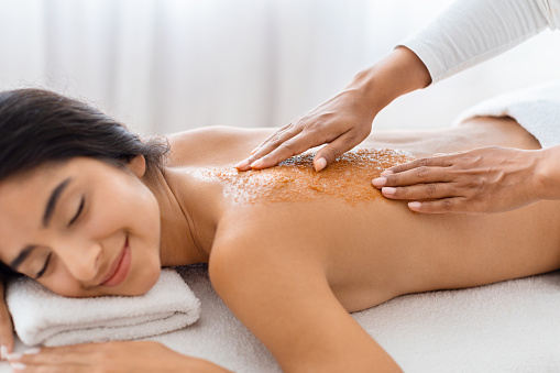 Serene relaxed young indian woman deeply relaxing as a spa professional applies an exfoliating treatment to her back, promoting wellness and skin health, closeup