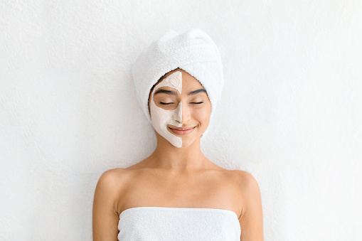 Relaxed young indian woman with a towel on her head and a half-applied facial mask, smiling with eyes closed in a blissful spa setting, white background, top view. Skin care treatment