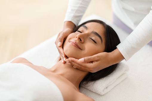 Beautiful young indian woman enjoying face lifting massage in spa salon, professional therapist making skin lift procedure to relaxed brunette lady lying on table in wellness center, closeup