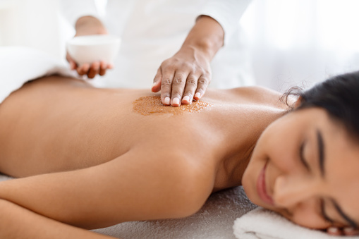 Closeup of joyful young indian lady having skin scrubbing procedure at spa salon. Therapist applying exfoliating body mask on sleeping eastern woman back at luxury spa, body care concept