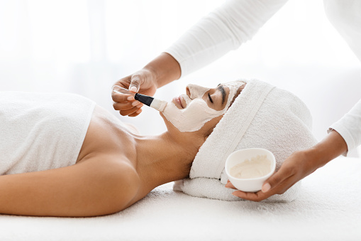 In a spa facility, a striking Indian woman is treated to a skincare session, with a skilled aesthetician administering a detoxifying mud mask on her elegant facial features