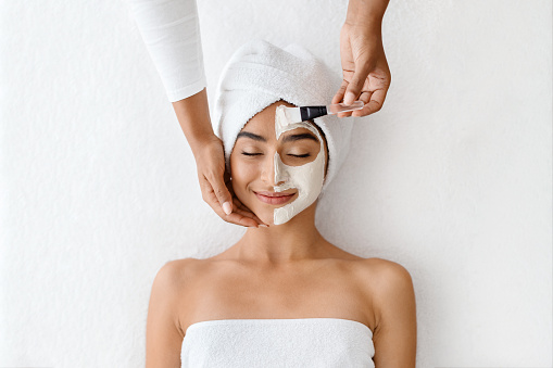 Anti-Aging Skincare. Cosmetologist applying clay mask on face of young indian woman, beautiful lady with towel on head lying on table, enjoying beauty tratments in spa salon, view above