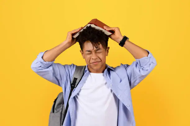 Overwhelmed young black man in casual outfit with backpack places red book on his head, eyes closed, showing signs of stress or frustration against yellow background