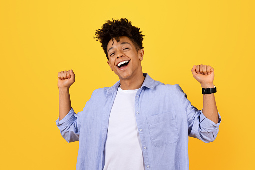 Ecstatic young black man with curly hair laughing and raising clenched fists in victorious or celebratory gesture, wearing casual blue shirt against vibrant yellow backdrop