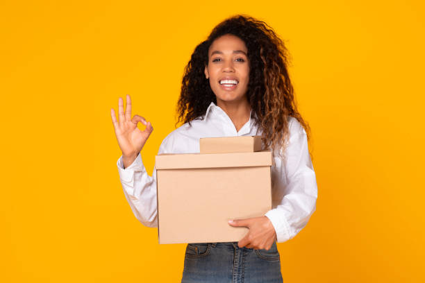 istock Black Woman Holding Parcel Box Gesturing Okay On Yellow Background 1996049444