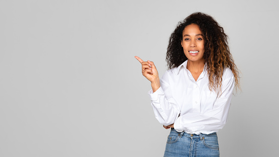 Positive Young Black Woman Pointing Finger Aside At Empty Space For Offer Advertisement, Smiling To Camera While Standing Over Gray Studio Background. Look There Banner, Panorama