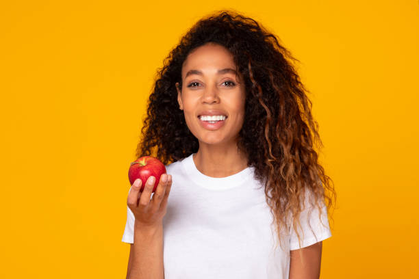 istock Cheerful black young lady holding fresh red apple, yellow backdrop 1996050517