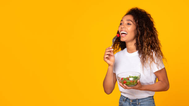 istock Black millennial lady happily eating vegetable salad against yellow backdrop 1996051058