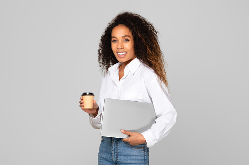 African American lady smiling holding her laptop computer, standing with paper coffee cup, in studio on gray background. Digital technologies, freelance and online career concept