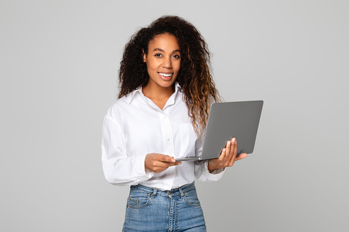 Young African American woman holding laptop browsing through modern websites, smiling to camera while posing with digital notebook device, in studio on gray background. Online tech concept