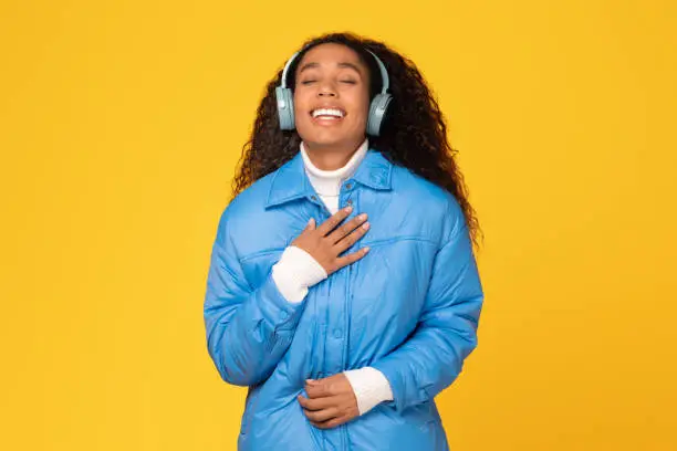 Young black woman wearing winter coat delights in music, singing with her eyes closed, against sunny yellow background, feeling the holiday cheer