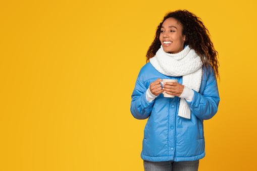 Joyful young black lady in winter jacket and scarf, holding cup of hot beverage, looking aside at free space on yellow background, holiday warmth in her eyes