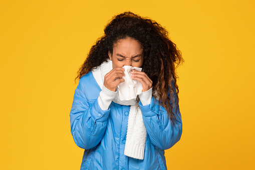 Young black lady in winter jacket and scarf blowing her nose into handkerchief against yellow background, depicting a seasonal cold scenario