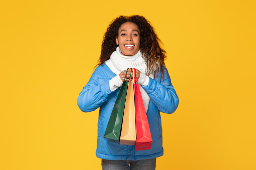 happy young black lady in winter attire holding shopper bags, enjoying seasonal sales, posing on bright yellow background and smiling at camera
