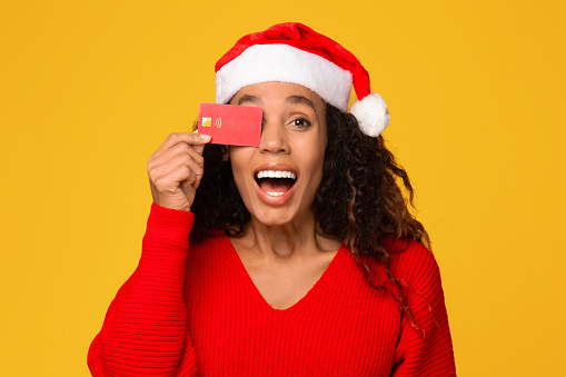 Portrait of young excited black lady, dressed in Santa hat, playfully covering one eye with credit card, ready for Christmas sales on yellow backdrop