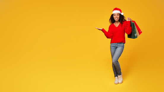 Enthusiastic black lady in Santa hat, gripping shopping bags in hand, pointing at free space against vivid yellow background, embodies Christmas sales, full length, panorama