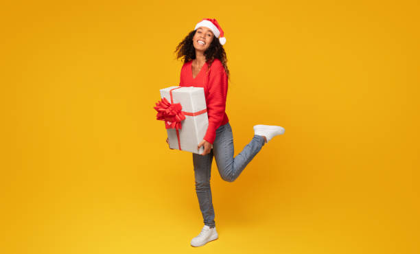 istock Black lady with Santa hat holding gift on yellow background, full length 1996067224
