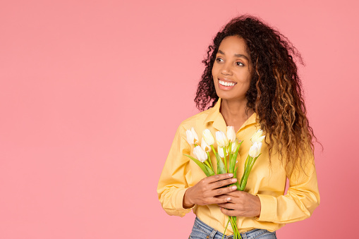 Happy black lady with curly hair, in blouse, holds bouquet of fresh white tulips against soft pink background, capturing the essence of spring and floral beauty, free space