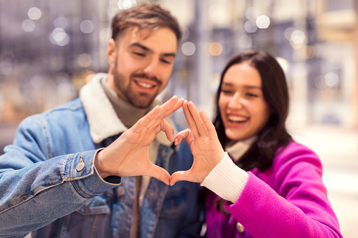 Young European couple romantically shapes heart with their fingers during winter evening walk in city. Man and woman, smiling immersed in holiday happiness during Christmas and Valentine's Day.