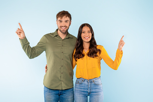Happy man and woman with bright smiles, standing side by side and pointing upwards, suggesting shared goal or vision, against light blue backdrop