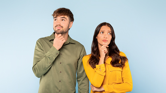 Pensive young man and woman in casual attire with hands on their chins looking upwards, both lost in thought on light blue background