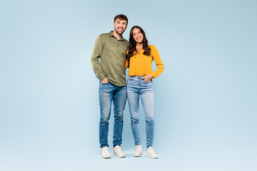 Happy couple standing close in denim jeans and colorful shirts, sharing a comfortable moment together, all set against calm blue background, full length