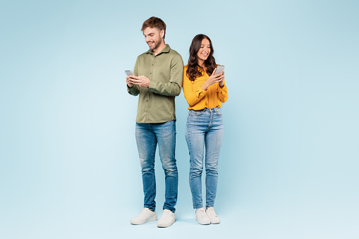 Contented couple each focused on their smartphones, texting and interacting with their digital worlds while standing side by side, against blue background, full length
