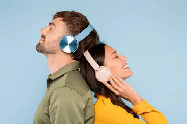 Smiling man in olive shirt and woman in mustard blouse with closed eyes, feeling the music in their headphones, back-to-back on a light blue backdrop