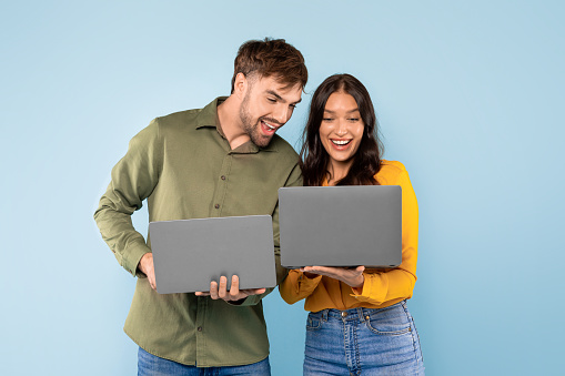 Cheerful young couple comparing notes on their laptops, sharing a laugh over a digital discovery against a bright blue background, surfing internet