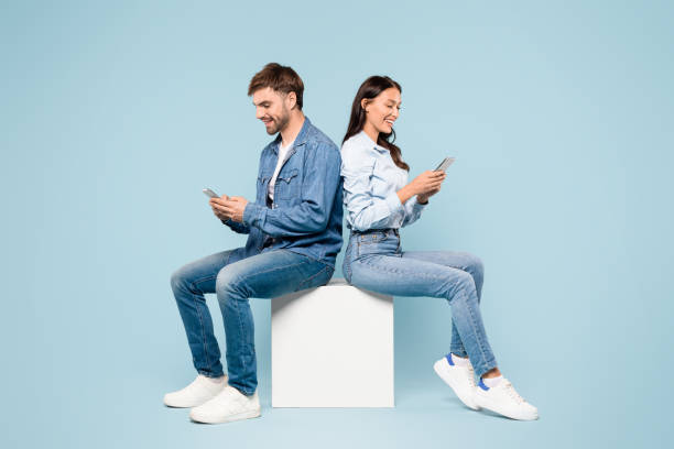 istock Man and woman in denim using smartphones back-to-back on a white cube, blue background 1996095663