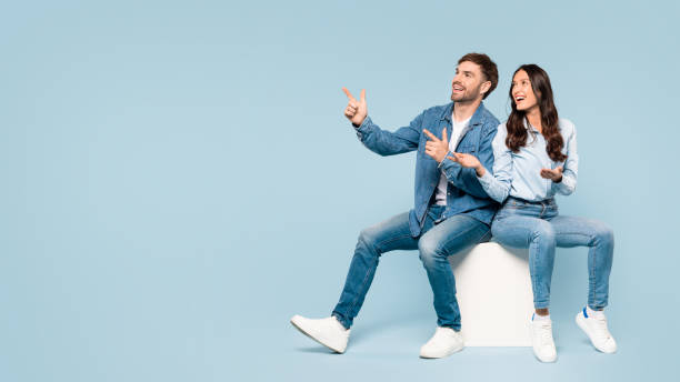 istock Playful couple in denim pointing outwards, seated on cube, free space 1996095667