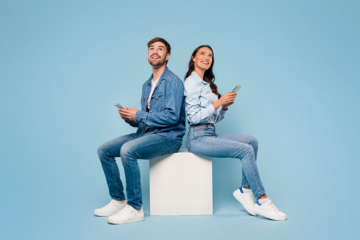 Seated back to back, young European man and woman use their mobile phones, smiling looking at free space on blue studio background, depicting relaxed connectivity