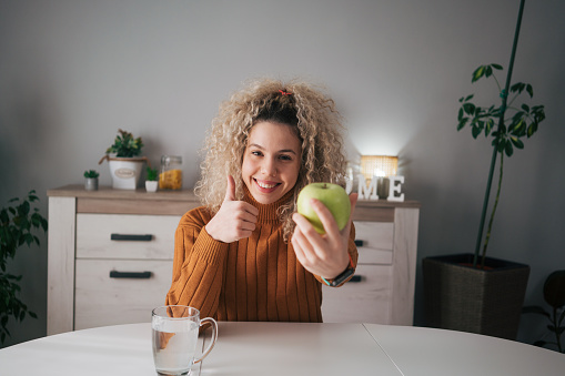 Beautiful woman with curly hair holding a green apple while sitting in the dining room,she gives a thumbs up