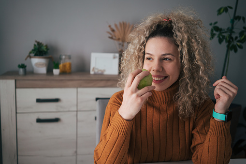 Beautiful woman with curly hair holding a green apple while sitting in the dining room