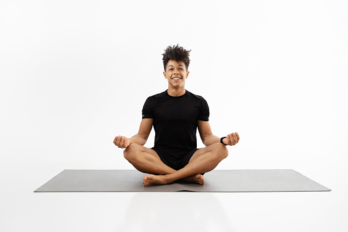 Young fitness guy in fitwear practices yoga sitting in lotus position on gym mat, meditating and relaxing on white studio background. Sporty man smiles and enjoys calmness during workout meditation