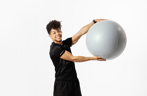 Smiling young latin guy lifting fitball smiling to camera while exercising on white studio background. Determined man in sports outfit practicing gymnastics, doing aerobic exercises