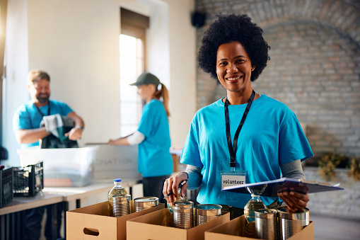 Happy African American woman going through donated supplies while volunteering at food back and looking at camera.