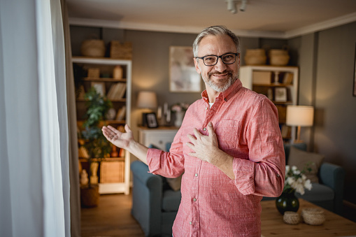 A mature man is cheerfully standing in his apartment and welcoming guests, with a cheerful expression on his face