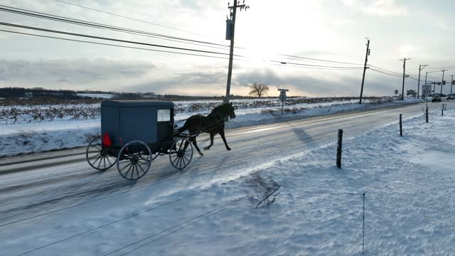 Amish buggy on snowy road in Lancaster County, PA, under a sunset sky. Aerial tracking shot as snow drifts on rural USA road.