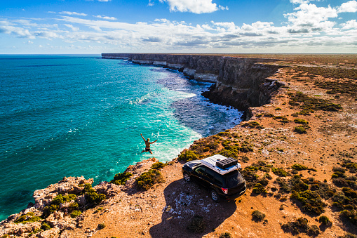 Aerial view of car parked with camper and happy young man jumping for joy over the Great Australian Bight. Captured while crossing the Nullarbor.