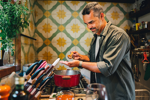 Smiling mature man using a spoon to taste food cooking in a pot on a stove in his kitchen at home