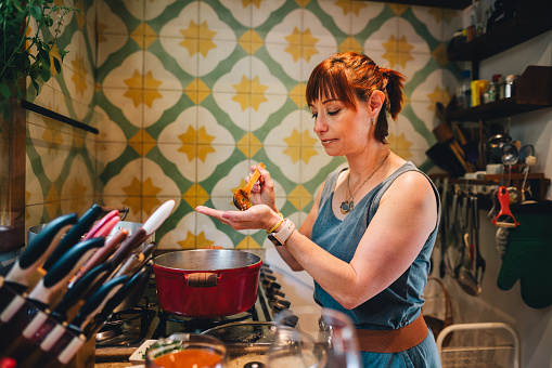 Smiling mature woman using a spoon to taste food cooking in a pot on a stove in her kitchen at home