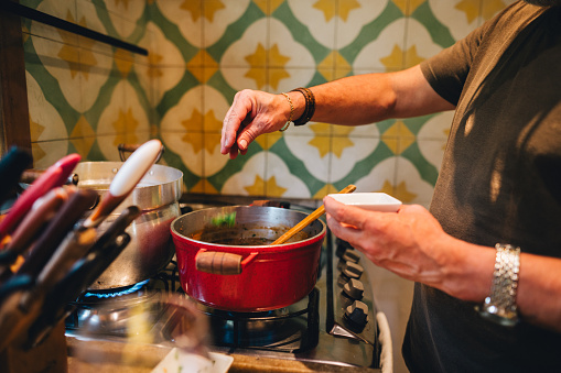 Close-up of a mature man seasoning a meal cooking in a pot on a stove in his kitchen at home