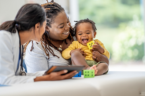 A young Mother, of African decent, brings in her toddler for a check-up.  She is holding the little girl gently on the exam table as she talks with the doctor who is holding out a tablet with test results on it.