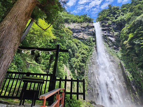 This image depicts a captivating scene of a towering waterfall cascading down a rocky cliff surrounded by lush greenery. In the foreground, there is a wooden viewing platform, which includes a bench for visitors to sit and admire the waterfall. The platform is secured with black railings and is accessible by steps with a red side railing. The waterfall appears to be in a forested area, and the sky is blue with a few wispy clouds, suggesting a fair weather day. The waterfall's mist can be seen dispersing in the air, adding to the dramatic and refreshing ambiance of the scene.