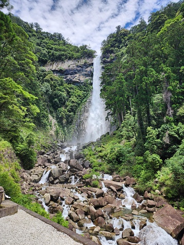 This is an image of a majestic waterfall cascading down a steep, rocky cliff surrounded by lush greenery. The waterfall appears to be quite tall, and the force of the water creates a mist near the base. The waterfall's powerful stream flows down into a rocky riverbed where the water continues its course, evident by the white rapids over the uneven stones. To the left in the foreground, there is a man-made path or viewpoint that provides a place for observers to take in the scenic beauty. The surrounding vegetation is vibrant and dense, indicating that this could be a location within a tropical or subtropical forest. The sky is partially visible at the top, hinting at fine weather with scattered clouds, which allows sunlight to illuminate the scenery, enhancing the natural beauty of the area.