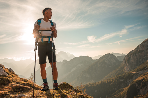 A mature Asian man enjoys a mountain hike on a sunny day, navigating the trail with sunglasses and hiking poles.